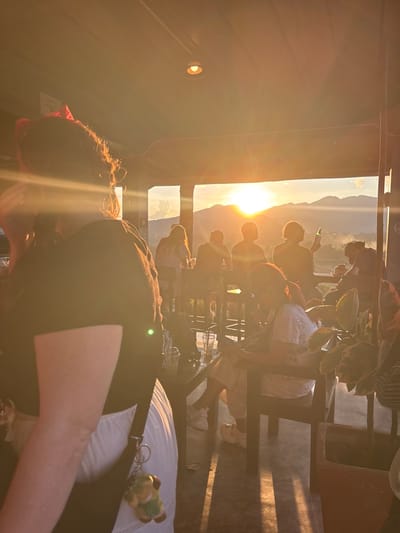 The image depicts an outdoor or semi-outdoor elevated establishment in Ban Mae Ping, Thailand, at sunset. The setting sun, positioned centrally on the horizon above a range of mountains, casts a strong golden light, silhouetting many subjects and creating significant lens flare. In the immediate foreground, the back and right profile of an individual with dark, curly hair adorned with a red bow is visible, largely silhouetted. This person wears a dark top and white lower garment, with a shoulder strap and a light-colored, animal-shaped keychain (yellowish-brown and green) attached.
Towards the midground, several other individuals are seated and standing. On the far right, a person is seated, looking downwards, possibly at a handheld device, wearing a white top and light-colored pants. To their left, another individual is seated, holding a green bottle. Other figures are visible further back, mostly as silhouettes against the bright horizon, some appearing to be seated at high-top tables or bars. Various clear plastic cups with straws are present on dark-colored tables. The establishment features a dark roof, visible pillars, and a ceiling light fixture. The ground is a light gray surface, exhibiting long, distinct shadows cast by the subjects and structures due to the low sun. In the far background, beyond the people and structures, a panoramic view of layered mountains is visible under a sky transitioning from intense orange-yellow at the horizon to lighter hues above. Potted plants with large leaves are visible in the right midground.