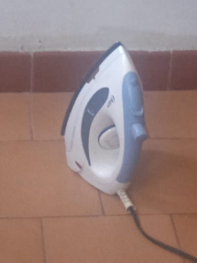 The image shows a white and blue steam iron resting on a tiled floor. The iron has a grey accent and a visible power cord. The background consists of a wall with reddish-brown tiles forming a lower border. The scene is likely indoors, given the tiled surfaces and wall. No people are visible, and there is no indication of specific time of day or weather conditions. The image's context is Puerto La Cruz, Venezuela. The word "Oster" is visible on the side of the iron. The activity depicted is simply the iron at rest.