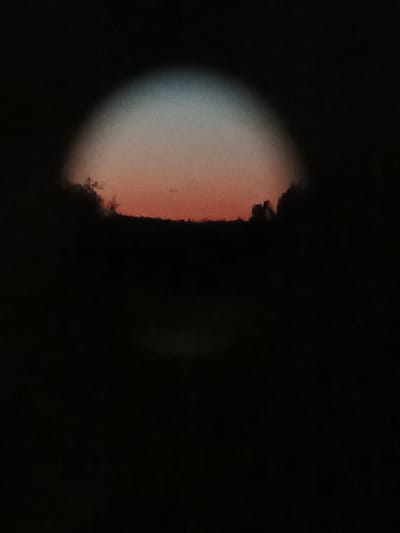 The image shows a dark, circular vignette of a sky at dusk. The sky within the vignette transitions from a pale blue at the top to a warm orange-red near the horizon. Silhouetted clouds or land formations are visible along the lower edge of the vignette, obscuring the lowest part of the sky. The majority of the image, outside of the central vignette, is completely black, suggesting the photograph was taken in very low light conditions. There are no discernible people, objects, or specific details about the environment, and no specific location in Воскресенское, Russia can be determined from the image.