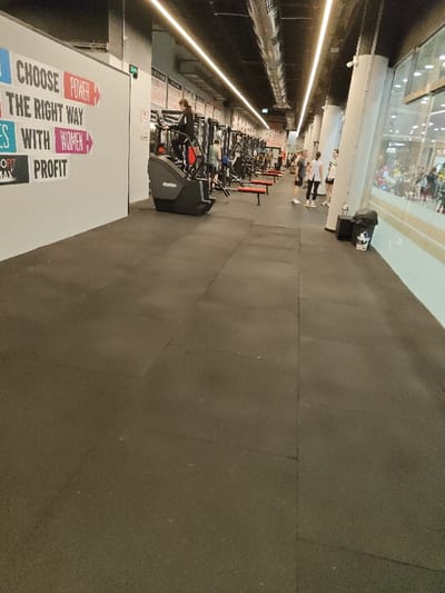 The image depicts a gym interior with workout equipment, including a stair climber. People are visible using the equipment and standing nearby. The gym has a long, narrow layout with bright overhead lighting. Inspirational phrases adorn the wall on the left. The flooring is dark and rubberized. Outside the gym's large windows, pedestrians are visible.