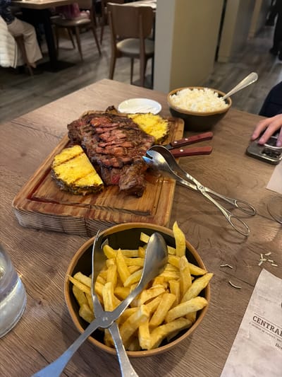 The image shows a table setting in a restaurant in São Mamede de Infesta, Portugal, featuring a wooden cutting board with grilled steak and pineapple, a bowl of rice, a bowl of fries, and serving utensils. A piece of paper with "CENTRAL" and "S. MAMEDE DE INFESTA, RIO T" printed on it is also visible. A person's hand holding a phone is seen in the upper left corner. The table is wooden. The background is slightly blurred, showing chairs and other restaurant elements.