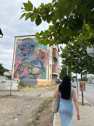 A woman with long, dark hair, wearing a light blue knitted dress and carrying a light grey tote bag, walks away from the camera down a sidewalk. To her left, a tall building features a large, colorful mural on its side depicting a silver teapot, a tray of figs, and intricate patterns. The mural is framed by a blue border. The building itself is multi-story with balconies. To the right of the woman is a street with cars and a sidewalk. A red and yellow sign is attached to a pole near the sidewalk. The environment is urban, with some greenery from trees and scattered debris on the ground. The sky is overcast. The building with the mural is located in Derbent, Russia.