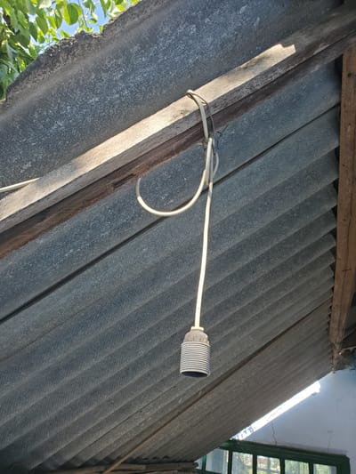 An exterior, low-angle shot captures a segment of an old, weathered corrugated roof and its supporting wooden beams. A bare light socket with a white cord hangs from a beam, attached by wire and a loop of the cord itself. The corrugated roofing material, likely asbestos cement, is gray and shows signs of age. Sunlight streams through gaps and illuminates the scene, casting shadows. Trees with green leaves are visible in the upper left corner, suggesting an outdoor setting. The lower right of the image reveals a portion of a building wall with cracks and a window with green trim, hinting at a rustic, possibly rural or older urban environment. The location is the city of Konstantinovka, Moldova.
