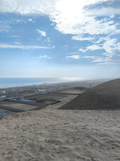 The image shows a view overlooking a landscape in Huanchaco, Peru. The foreground is dominated by a sandy, textured ground, likely a dune or hill. To the right, a darker, more substantial hill slopes down towards the developed area. In the middle ground, a grid-like pattern of buildings and fenced enclosures is visible, extending towards the beach and the ocean. The buildings are primarily low-lying, simple structures. The beach stretches along the coastline, with visible waves. The sky is predominantly blue with scattered clouds, and the sun is bright.