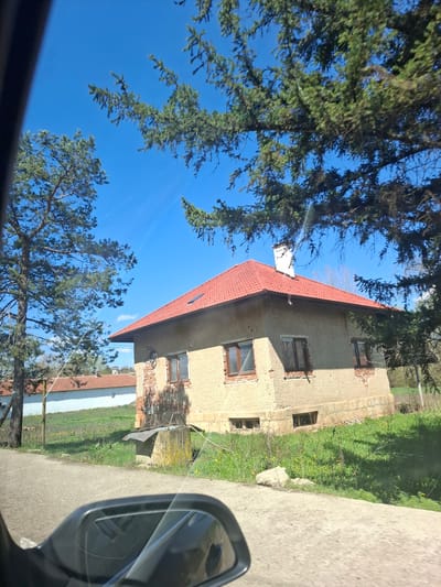 The image shows a building with a red tiled roof, possibly residential, viewed from a car window. Part of a side mirror is visible. The building appears to be in a state of disrepair, with some brickwork exposed. There are trees in the foreground, obscuring the sky. The location is Studeno Buche, Bulgaria, based on prior context. The road and vegetation adjacent to the building are visible. No people are identifiable in the image.