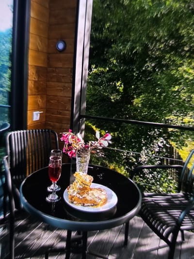 The image depicts an outdoor seating area with a table and two chairs, set against a backdrop of lush green trees. On the black, circular table, there are two glasses filled with red liquid, a vase containing pink and white flowers, and a plate with a slice of cake. The table is situated on a wooden deck. Behind the table and chairs, a wooden-paneled wall features a light fixture and an electrical outlet. The scene is bathed in sunlight, casting shadows on the deck. The general impression is that of a peaceful outdoor setting, likely a balcony or patio, with a refreshing drink and a slice of cake. The specific location in Dubno, Ukraine, is not discernible from the image itself.