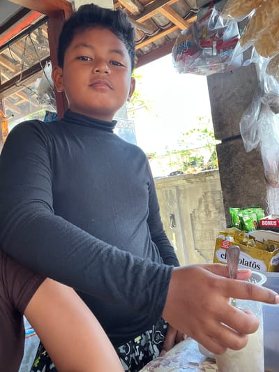 The image shows a young boy in Mendoyo, Indonesia, looking up at the camera with a neutral expression. He is wearing a black long-sleeved shirt. His right hand, holding a spoon, is raised near his face. In the background, there are various packaged snacks, some in plastic bags. One visible package has text that reads "BONUS," "MAKIN," "RENYAH," "MAKIN," and "LEZATO." The scene appears to be outdoors, possibly at a market or roadside stall, with natural light suggesting daytime. The overall setting seems casual and everyday.