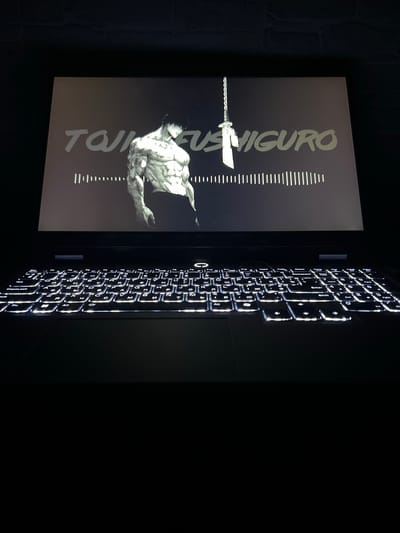 A laptop is positioned on a dark surface, likely a desk or table, with its screen illuminated. The screen displays an image of a muscular, tattooed male figure in a dark, stylized artwork, accompanied by text that appears to read "TOJI FUSHIGURO" in large, light-colored lettering. Below the text and figure, a waveform or equalizer graphic is visible. The laptop's keyboard is also lit up, revealing its keys. The background is dark, with a hint of a textured surface, possibly brick or stone, visible at the top. The overall scene is dimly lit, with the primary light source being the laptop screen and keyboard. There is no indication of a specific location in Sobishchytsi, Ukraine, or any other city.