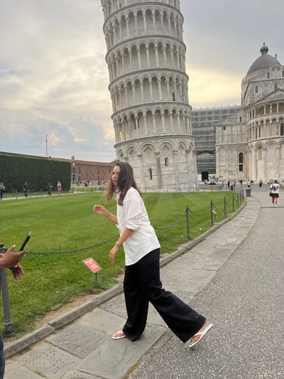 A photograph shows a woman pretending to hold up the Leaning Tower of Pisa. She is standing on the sidewalk beside a grassy area enclosed by a chain fence. The tower is behind her to the left, and the Pisa Cathedral is further back. There are other tourists visible. The sky is cloudy. Someone is holding a phone to take the photograph. The location is Pisa, Italy.