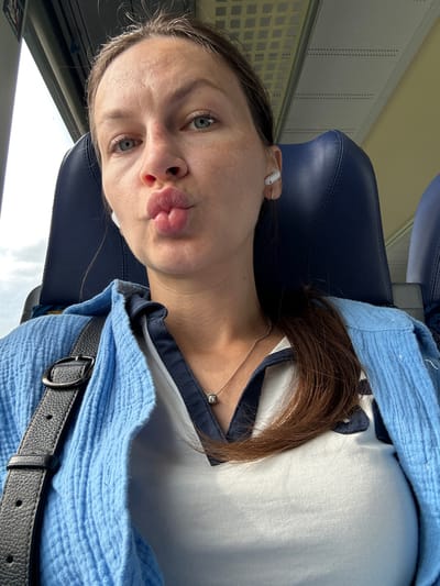 A fair-skinned female, approximately in her 20s or 30s, is captured in a close-up selfie within what appears to be a public transport vehicle. She has light blue eyes, visible freckles on her nose and cheeks, and her lips are pursed in a 'kissing' expression. Her dark hair is mostly pulled back, with some strands visible around her face. She wears white wireless earbuds in both ears. A thin silver necklace with a small, dark, square-shaped pendant is visible around her neck.
She is dressed in a light blue, textured, long-sleeved shirt or jacket, worn open over a white polo-style shirt with a dark blue collar. A black, textured leather strap, likely from a bag, is visible across her left shoulder. She is seated in a dark blue upholstered seat with a prominent headrest.
To her upper left, a window shows a bright, overcast sky. Partial German text, reading "ALKOHOLFREI" (alcohol-free), is visible on a white panel near the window. The ceiling of the vehicle is light-colored, featuring a grid pattern of small square vents on the left and horizontal dotted lines (possibly ventilation or speaker grilles) extending across.
The presence of the German text confirms the location is within Germany or a German-speaking region. However, there are no specific visual identifiers within the image, such as landmarks, specific vehicle markings, or unique architectural features, to definitively confirm the exact city as Oberndorf.