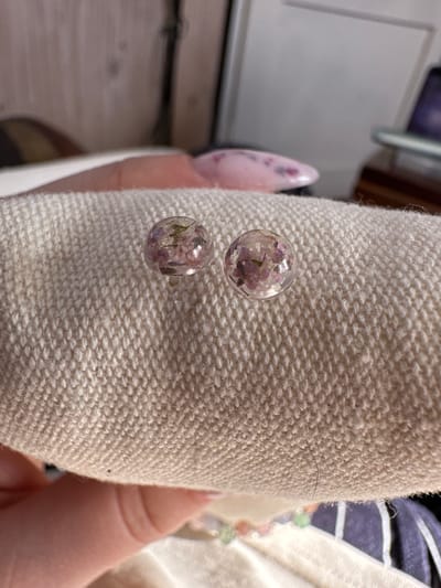 The image shows two round, clear earrings containing small, dried purple and green flowers resting on a textured, off-white fabric. A fingernail with pink nail polish and purple floral design is visible on the left, and part of a hand is seen on the right. The background is blurred, showing a white cabinet and a partially visible object with striped patterns. The image was not taken in Galiche, Bulgaria.