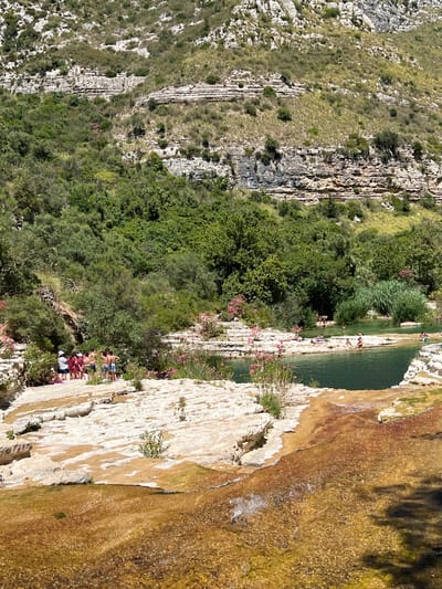 The image depicts a natural landscape featuring a river with clear, greenish-blue water flowing over textured, light-colored rocks. Several people are visible relaxing and swimming in the river and on the surrounding rocky terrain. Lush green trees and vegetation cover the hillsides, with rocky formations and striations visible on the slopes in the background. The scene appears to be a popular outdoor recreational spot, possibly within the city of Noto, Italy, known for its natural beauty. The water is shallow in some areas, revealing the rocky riverbed, while other parts are deeper and more shaded by the surrounding foliage. The overall impression is one of a serene and scenic natural environment with human activity integrated into the landscape.