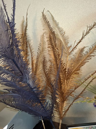 Two artificial plumes, one dark purple and one gold, are positioned vertically against a textured off-white wall. The plumes are feathery and branched, with fine, feather-like projections extending from their central stems. The gold plume is positioned slightly behind and to the right of the purple plume. The lighting creates shadows from the plumes on the wall. In the lower left corner, a reflective surface, possibly a table or shelf, is visible, along with a dark object. In the lower right corner, the edge of a framed picture is visible. The image is a close-up, top-down shot of these decorative plumes.