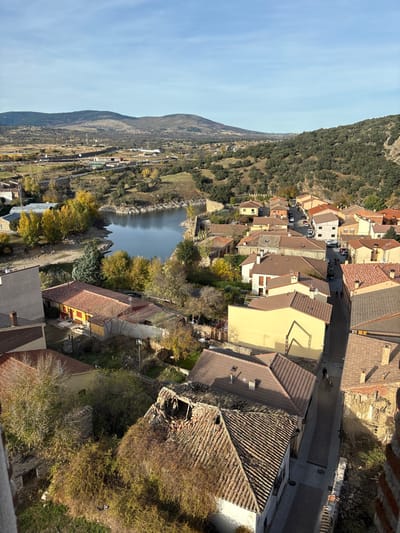This elevated, forensic-style image captures a portion of the town of Buitrago del Lozoya, Spain, under clear, sunny conditions. The scene primarily depicts residential architecture, a body of water, and surrounding natural landscapes.
In the foreground and mid-ground, a dense cluster of buildings with predominantly reddish-brown terracotta tiled roofs is visible. Building facades exhibit varying colors, including beige, yellow, and white. A prominent detail in the lower-mid foreground is a roof with significant structural damage, featuring a large, irregular collapse exposing internal timber framing and debris. Adjacent to this, another roof is extensively overgrown with dense, dried brown vegetation. Narrow, dark-paved streets wind between these structures.
Several parked vehicles are visible on streets in the mid-ground. At least two individuals are discernible walking along a narrow street in the lower-right portion of the image, moving away from the viewer.
To the left and center-mid ground, a calm body of water, likely the Lozoya River or an associated reservoir, is present, reflecting the blue sky. Its banks reveal areas of exposed earth, and stone walls, possibly historical fortifications, line sections of the waterway. Trees with bright yellow autumnal foliage are interspersed throughout the town and along the water's edge.
The background consists of rolling hills and mountains, largely covered with varying densities of green and brown vegetation, extending under a clear blue sky with minimal cloud cover. Distant structures and sparse development are visible in the expansive landscape beyond the town. The viewpoint appears to be from an elevated structure, with a partial brick wall visible in the extreme lower right.