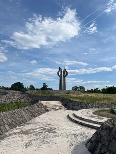 The image shows a monument located in Barutana, Montenegro. The monument features three stylized, outstretched arms reaching towards a cloudy sky. The base of the monument is surrounded by stone walls and landscaping. A set of curved steps leads up to the monument area.