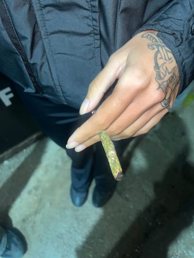 A person is holding a lit marijuana cigarette in their left hand. The person is wearing a black jacket and has a tattoo on the back of their hand. They appear to be sitting. The background is an out-of-focus concrete surface. It is not possible to determine if this photo was taken in Lomas de Zamora, Argentina or any specific location.