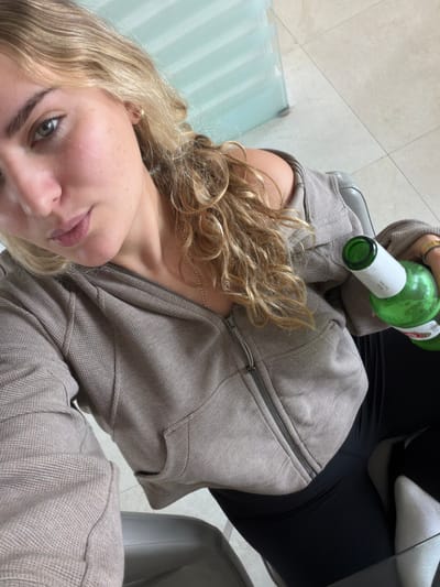 A young woman with blonde, curly hair and freckles is seated and angled towards the camera, holding a green glass bottle. She is wearing a brown zip-up hoodie over black leggings. The setting is indoors, possibly a room in a residence or establishment in Key Biscayne, United States of America, evident by the tile flooring and hint of a glass table or wall. The floor is beige tile and there is a glimpse of a chair.