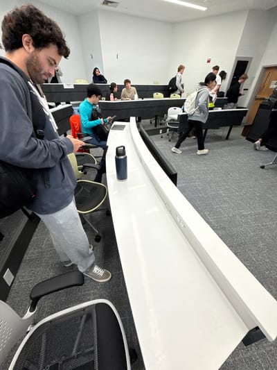 An indoor scene within a modern, tiered lecture hall or classroom in Wellesley, United States, is depicted. The room features white walls, a grey patterned carpet, and curved black and white desks with integrated power outlets along their top surfaces. Black swivel chairs are positioned at each desk.

In the immediate foreground, a male with dark, curly hair and a beard, wearing a dark grey hoodie, light grey sweatpants, and grey sneakers with white laces, stands. He is looking down at a small electronic device held in both hands. A black shoulder bag is slung over his left shoulder. To his right, on the white desk surface, a dark blue insulated water bottle with a black handle is positioned.

Further into the mid-ground on the left, another male with dark hair, wearing a bright turquoise long-sleeved shirt, sits at a desk, looking down at a dark electronic device in his hands. Behind him, several other individuals are seated at desks, some facing forward, others looking to the side.

On the right side of the mid-ground, a male with light-colored hair, wearing a light grey hoodie and a white backpack, walks from right to left, partially obscured. Other individuals are visible standing and walking in the background near a light brown wooden door with a window and a handle, located on the far right. The overall activity suggests a transition period, possibly before or after a class.
