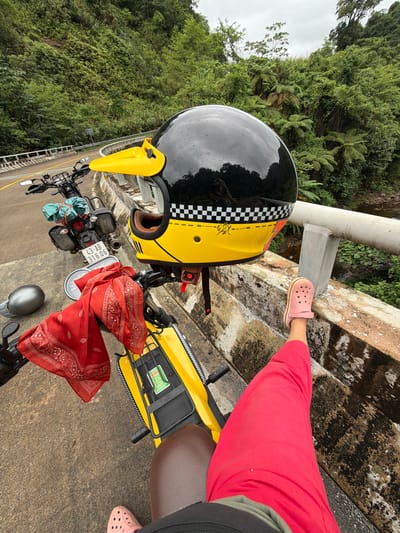 A person's legs, clad in bright red pants and pink Crocs, are propped up on a concrete bridge railing. The perspective suggests the photo is taken from a seated position on a yellow motorcycle. A black and yellow motorcycle helmet with a checkered pattern rests on the handlebars, partially obscuring the rider's view of the road ahead, which curves to the left. A red bandana is draped over the handlebars, and a silver helmet lies on the ground. The motorcycle is parked on a road that runs alongside a lush, green hillside covered in dense vegetation. A river or stream flows below the bridge. The scene appears to be in a rural area, possibly within Thượng Trạch Commune, Vietnam, given the surrounding foliage and the style of the motorcycle and road. The sky is overcast, suggesting a humid climate.