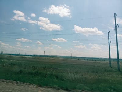 The image depicts a vast, open field under a partially cloudy blue sky. Several power lines stretch horizontally across the frame, supported by utility poles, two of which are prominent in the foreground on the right. The field itself is covered in green grass, with patches of lighter, dry grass or vegetation interspersed. In the distance, a treeline is visible, marking the horizon. The scene appears to be rural, suggesting a location outside of a dense urban area. The image was taken during daylight, likely in the daytime.
