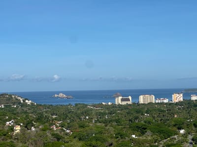 This is a high-angle, wide-shot, daytime photograph of Ixtapa Zihuatanejo, Mexico, showcasing a coastal landscape. The upper portion of the image is dominated by a clear blue sky with a few faint white clouds and two circular smudges that appear to be lens flares. Below the sky, a wide expanse of deep blue ocean stretches across the horizon, dotted with several rocky outcrops and small islands.

The foreground and midground are filled with lush green vegetation, primarily palm trees and other dense foliage, characteristic of a tropical environment. Interspersed within the greenery are numerous residential buildings and several prominent, modern hotels. These structures vary in size and architectural style, with some being multi-story resorts and others appearing to be smaller villas or housing complexes.

On the left side of the image, a hillside is covered with white-roofed houses, cascading down towards the ocean. In the center and right, several large hotels with light-colored facades stand out against the natural landscape. One prominent hotel, a tall, cream-colored building, is situated on the right side of the midground, facing the ocean. Further to the right, other hotels and buildings are visible along the coastline. A winding road is discernible in the lower right quadrant, disappearing into the dense vegetation. The scene depicts a developed coastal resort area integrated with its natural surroundings. There are no people or animals clearly visible in the image.