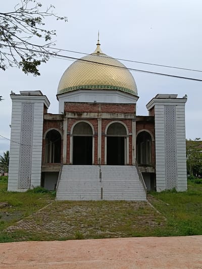 The image shows a mosque under construction in Meureudu, Indonesia. The mosque features a prominent golden dome with intricate green detailing around its base. The main structure is built with exposed red bricks and has large arched entrances. On either side of the entrance, there are tall, white pillars decorated with a repeating geometric pattern. A wide, white staircase leads up to the main entrance. The surrounding area is covered in grass and some unpaved ground, with a few trees visible in the background. The sky is overcast. There are no people visible in the image. The scene suggests ongoing construction work, with some elements of the building completed while others remain unfinished. The location appears to be a semi-urban or rural setting, given the vegetation and the general unfinished state of the grounds.