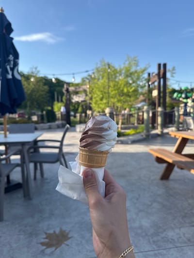 The image, captured in Chelsea, Canada, depicts a person's hand holding a swirled vanilla and chocolate soft-serve ice cream cone on a bright, sunny day. The ice cream is served in a light brown waffle cone, with a white paper napkin wrapped around its base. A gold chain bracelet adorns the wrist of the hand holding the treat.

The scene is an outdoor setting, likely a patio or eating area. The ground is a light gray concrete, and a distinct, dry leaf shape is visible on it to the right. In the mid-ground, there are dark metal chairs and tables, along with a wooden picnic table to the upper right. The background features lush green trees and what appears to be part of a building or complex with wooden structures. String lights are strung across the scene, suggesting an inviting outdoor space.

The sky is a clear, vibrant blue, indicating fair weather and daytime hours. A dark blue umbrella, partially visible in the bottom left corner, shows some white, illegible text or logos. The overall atmosphere conveys a sense of relaxation and enjoyment, typical of an outdoor leisure activity.