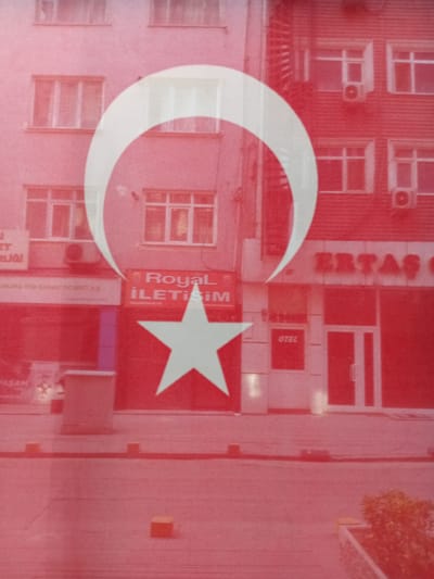 The image shows a building facade partially obscured by a large Turkish flag. The building has multiple windows and some visible air conditioning units. There are signs on the building, including "Royal İletişim" and "OTEL". The Turkish flag, with its white crescent and star on a red background, dominates the foreground and reflects onto the building. The scene appears to be a street in Kırıkkale, Turkey, given the presence of the Turkish flag and typical urban architecture. The image quality is slightly blurred, suggesting it may have been taken through a window or a reflective surface. There are no people visible in the image.