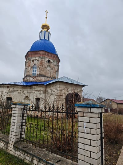 The image shows an old, weathered church in посёлок совхоза "Буденновец", Russia. The church has a blue dome and a golden cross. The walls of the church are made of white brick, and the building is surrounded by a black iron fence with white brick pillars. There are bushes and grass inside and outside the fence. In the background, there are residential buildings and an overcast sky.