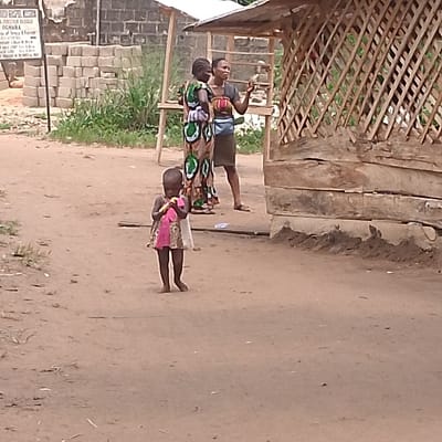 The image shows a young girl wearing a pink dress holding a yellow object in her hands, standing in front of a wooden shack with a thatched roof in Adanisievbe, Nigeria. Two women are standing behind the young girl. One is wearing a green and white dress and the other is wearing a blue dress. The woman in the green and white dress is looking down at the young girl and appears to be talking to her. The woman in the blue dress appears to be pointing to something off-camera. The scene is set against a backdrop of a dirt road and a green bush. The shack is made of wood and has a thatched roof. There are some concrete blocks on the ground near the shack. The image appears to have been taken on a sunny day. The young girl is smiling and appears to be happy. The location is 5.955, 5.703 in Adanisievbe, Nigeria. The image was likely taken outside of the shack on a dirt road. There is no sign of any traffic or other people in the immediate vicinity.