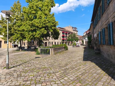 The image depicts a sunlit cobblestone plaza in Erlangen, Germany, on a clear day with a few scattered clouds. In the foreground, a modern street lamp casts a long shadow across the uneven, moss-interspersed cobblestones. To the left, a large, leafy tree with yellow blossoms dominates the scene, its branches extending towards the sky. Behind the tree, a building with large windows and a red awning is visible.
The plaza is bordered by traditional stone buildings with shuttered windows, typical of European architecture. The building on the right, in the foreground, features rows of blue shutters and a stone facade. In the mid-ground, a low stone wall with a metal railing and a gate separates the plaza from a grassy area. A bike rack is attached to the wall, with several bicycles parked nearby. Beyond this, more buildings with red awnings and balconies are visible, and a few people are seated at tables outside a cafe. In the distance, the cityscape of Erlangen is hinted at with more multi-story buildings. A dark-colored car is parked on the left side of the plaza. The overall atmosphere is peaceful and picturesque, suggesting a pleasant day in a historic European city.
