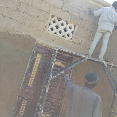 The image depicts an outdoor construction or renovation scene in Dan Maliki, Nigeria, featuring two male individuals and a metal scaffolding structure against a building wall. The wall is constructed of light-colored concrete blocks, partially covered with rough, light brown plaster. A white decorative ventilation block, featuring an '8' shape pattern, is embedded in the wall above a dark brown double metal door adorned with gold-colored ornate vertical and circular designs.
One individual, wearing a light-colored long-sleeved shirt and light-colored jeans, stands on the upper platform of the metal scaffolding. Their left hand is positioned against the concrete block wall, consistent with applying or inspecting plaster. The second individual, viewed from behind, wears a grey long-sleeved garment and a patterned cap. Their right arm is extended upwards, grasping a vertical bar of the scaffolding structure, indicating support or stabilization. The scaffolding is a rudimentary dark-colored metal structure with visible wear and light paint splatters. The scene is illuminated by natural light, suggesting a daytime setting.