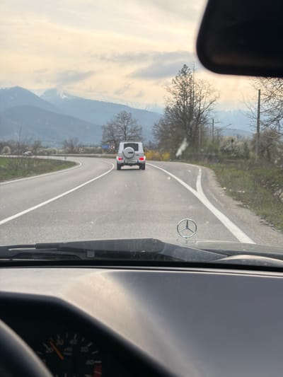 A vehicle is driving on a paved road with a white dividing line. The road appears to be winding through a mountainous region. In the distance, another vehicle, possibly a Jeep or SUV, is visible on the same road. The scene is viewed from inside a car, with a partial view of the dashboard and steering wheel. A Mercedes-Benz emblem is visible on the windshield. Bare trees and some greenery line the roadside. Mountains are visible in the distance, suggesting a location in a hilly or mountainous area. There is no indication that the photo was taken in Mezdreya, Bulgaria.