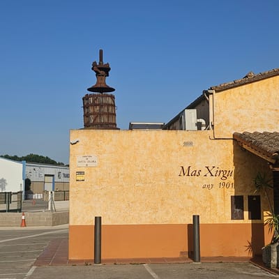 A close-up, eye-level shot captures a rustic, weathered wine press displayed on the exterior of a building. The wine press, made of wood and rusted metal, sits atop a textured, ochre-colored wall. To the right, the building's facade is a warm yellow with a red-tiled roof. Text on the wall reads "Mas Xirgu" and "any 1901". A white sign with blue text "CARRETERA DE SANTA COLOMA" is affixed to the wall. In the background, industrial buildings and a fence are visible, along with a clear blue sky. The scene suggests a warm, sunny day, possibly late morning or early afternoon, given the light and shadows. The overall impression is one of historical charm mixed with a functional, possibly commercial, setting.