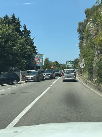 The image shows a traffic scene on a sunny day in Vrijesno, Montenegro. A line of cars is driving on a two-lane road, with a steep, vegetated rocky cliff face on the right side of the road. A speed limit sign indicating 40 is visible on the right. On the left side of the road, there are trees and a billboard advertising a beverage. The road has white lane markings. The sky is clear and blue. No people are clearly visible in the image, and the cars appear to be moving or stopped in traffic.