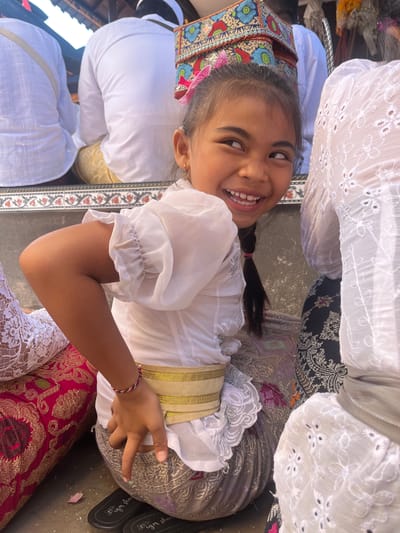A young girl with dark hair styled in a braid and a pink bow smiles and looks to her right while sitting on the ground. She wears a white blouse with ruffled sleeves and a gold sash. She is surrounded by people in white clothing, and colorful textiles are visible on the ground. The scene appears to be outdoors, possibly during a cultural event or ceremony. Based on the attire and the presence of what looks like traditional Indonesian religious adornments, it is likely this image was taken in Mendoyo, Bali, Indonesia.