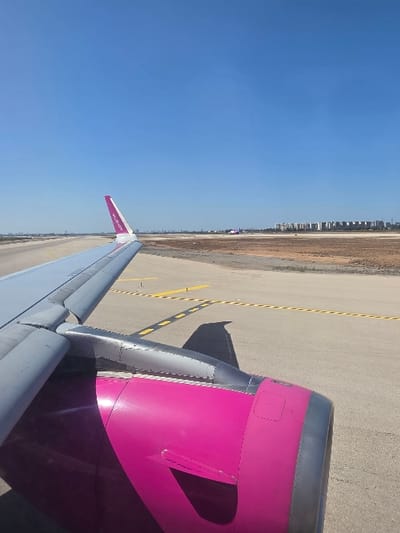 A forensic examination of the image reveals a detailed perspective from an aircraft window at an airport, likely Ben Gurion Airport (TLV), which borders the city of Yehud Monosson, Israel. The primary subject is the right wing and engine of a commercial passenger aircraft. The wing's upper surface is light gray, with visible leading-edge slats and sections of the flap system. The engine nacelle is a distinctive bright magenta (pink) color, with "WIZZ" prominently displayed on the aircraft's wingtip. The engine's intake lip and rear section are metallic silver/gray. A dark shadow of the wing and engine is cast onto the concrete/asphalt tarmac below, indicating bright, direct sunlight.

The aircraft is positioned on an airport surface, characterized by light gray concrete or asphalt with yellow painted lines and black markings, consistent with taxiway or apron designations. In the middle ground, beyond a strip of dry, light-brown earth, another aircraft, also featuring a magenta livery (suggesting another Wizz Air plane), is visible on a parallel taxiway or runway. In the far distance, a low-rise urban skyline is discernible against the clear, unblemished blue sky. This skyline consists of multiple multi-story residential or commercial buildings, consistent with a city like Yehud Monosson, Lod, or Ramla, as viewed from the vicinity of Ben Gurion Airport. No people are visible in the frame. The scene depicts an aircraft either stationary or in the process of taxiing on an airport ground.