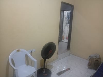 The image shows an interior scene within a room. On the left side, there is a white plastic chair with armrests, positioned against a light yellow wall. A white electrical outlet with a black cord plugged into it is mounted on the wall behind the chair. To the right of the chair, a black oscillating fan stands on a circular base. A broom with a red band around its bristles lies on the tiled floor in front of the fan. Further to the right, a beige backpack is placed on the floor, with a light-colored piece of fabric draped over its top. An open doorway is visible in the background, leading to another area with a window that has a metal grate. The floor throughout the room is tiled with a white and grey checkered pattern. The scene appears to be a residential interior. The specific location in Ekosa, Nigeria, is not determinable from the visual information provided.