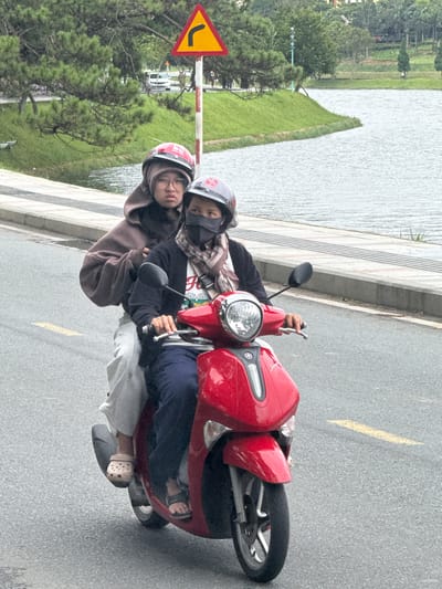 Two individuals, both wearing helmets, are riding a red scooter on a paved road. The person in front, presumably the driver, is wearing a black sweatshirt, a scarf, and a black face mask. The person behind them is wearing a brown hoodie and glasses. Both individuals are facing forward. The scooter has a visible headlight and mirrors. The background features a body of water, a grassy embankment, trees, and a traffic sign indicating a curve to the right. The scene appears to be in the city of Xuan Huong, Da Lat, Vietnam, given the presence of the lake and lush greenery characteristic of the area. The road surface is asphalt and shows yellow lane markings.