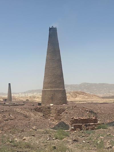 The image depicts a tall, tapering brick kiln or chimney, actively emitting smoke from its top against a clear, pale blue sky. The kiln is constructed from a dark, earthy material, possibly bricks or concrete, and stands prominently in the foreground, slightly to the left. Further in the mid-ground, another smaller, similarly shaped structure is visible.
The surrounding landscape is arid and rugged, characterized by dry, loose earth and rocky terrain. Sparse patches of green grass are visible on the ground in various areas. In the background, hazy, light-colored hills or mountains stretch across the horizon. Piles of what appear to be stacked bricks or construction debris are scattered around the base of the main kiln and further into the scene.
The activity shown is the active operation of the main kiln, indicated by the smoke plume. The scene suggests an industrial or semi-industrial setting, likely a brick-making facility. The time of day appears to be daytime under a clear sky, with a slight haze common in dry environments. No visible text is present in the image. The overall setting is consistent with arid regions where traditional brick kilns are common, aligning with the provided context of Pakistan.