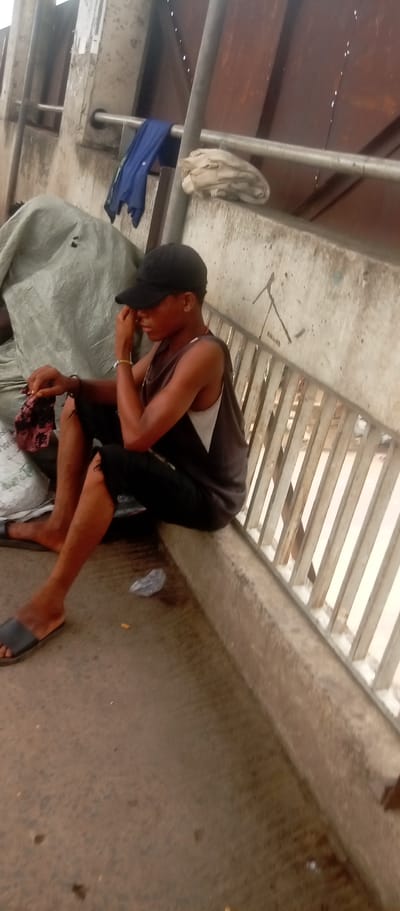 The image shows a young man sitting on a ledge near a metal railing in Idemili, Nigeria. He is wearing a black baseball cap, a tank top, and dark-colored shorts. He is wearing sandals. His left hand touches his face. He is sitting next to a bag. Discarded trash lies on the ground. Other items, including clothing, sit on a higher ledge.