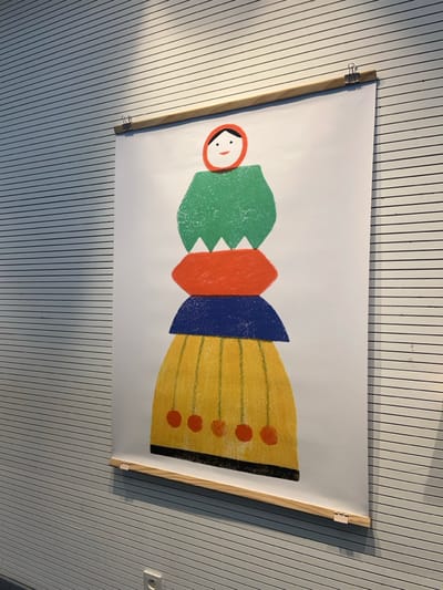 The scene depicts a vertically oriented poster mounted on a horizontally ridged, light gray wall in Sélestat, France. The poster features a stylized, abstract figure composed of stacked, textured, colorful shapes. From top to bottom, the figure includes a red circular head with simple black eyes and a red curved mouth, a green irregular form with a jagged lower edge, an orange flattened hexagonal shape, a dark blue curved half-moon shape, and a yellow bell-shaped form with vertical lines and five reddish-orange circular elements suspended from its base. The poster is secured at its top and bottom edges by light-colored wooden bars, clamped to the paper by three silver-toned binder clips on each bar. A diagonal shadow is cast across the upper left portion of the wall and the poster. A white electrical outlet with two circular receptacles is visible on the wall in the lower left quadrant. The precise building or establishment within Sélestat cannot be ascertained from the visual evidence.