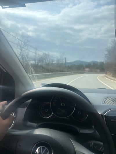 The image presents a first-person perspective from the driver's seat of a Volkswagen vehicle, identified by its steering wheel logo. A human hand is visible gripping the left side of the black steering wheel. The vehicle's instrument cluster is partially visible, indicating a speed of approximately 58 km/h. The windshield exhibits a dusty or streaked appearance.
Outside the vehicle, a two-lane asphalt road, marked with a solid white center line and a solid white line on the right shoulder, curves gradually to the right. Bare deciduous trees line both sides of the road, suggesting a late winter or early spring season. In the mid-distance, a tall, slender industrial-style tower or smokestack is visible on the left, rising above the treeline. Distant hills or low mountains form the horizon under a sky with scattered white and grey clouds. This scene is situated along a road in the area of Lukovo, Bulgaria.