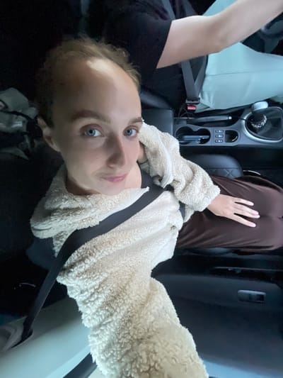 A person with blue eyes and light brown hair is sitting in the passenger seat of a car, looking at the camera. They are wearing a cream-colored fuzzy jacket over a dark top, and a black seatbelt is fastened across their chest. Their right hand is resting on their lap. The car interior is visible, including a gear shift, cup holders, and a dashboard with buttons. Another person's arm and shoulder, wearing a black t-shirt, are visible in the driver's seat. The scene appears to be a nighttime interior shot of a vehicle. There is no information in the image or provided context to indicate a location in Meshcheryaki, Russia.