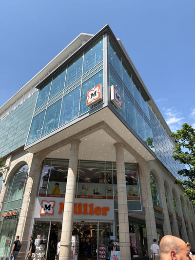 This is a vertical, low-angle shot of the exterior of a multi-story building, likely a department store, in Lörrach, Germany. The building features a modern glass facade on its upper levels, angled towards the sky, and a more traditional stone facade with arched windows on the lower levels. The sky is a clear, bright blue with a few wisps of clouds, suggesting a sunny day.

Several signs are visible on the building, including "Müller" in large orange lettering and the "Müller" logo, which appears to be a stylized flower shape with an "M" in the center. Other visible text includes "PARFUMERIE," "DROGERIE," "MULTI-MEDIA," and "SCHREIBWAREN," indicating the various departments or types of stores within the building. Some window displays are visible, showing products and advertising.

There are a few people visible in the scene, primarily on the sidewalk in front of the store. One person, a man with a shaved head, is prominently featured in the foreground, looking to the right and slightly away from the camera. Other individuals are seen walking or standing near the entrance. The overall atmosphere appears to be that of a busy commercial area on a pleasant day. The time of day is likely daytime, given the bright sunlight and clear sky.
