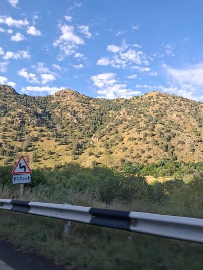 The image captures a view of a mountainous landscape under a partly cloudy blue sky. In the foreground, a road barrier with black and white stripes is visible, along with some vegetation. A triangular road sign with a black wavy arrow symbol and the text "↑1.7km↑" is mounted on a pole to the left of the road. The background features rolling hills covered with sparse vegetation and scattered trees, indicative of a rural or mountainous terrain. The scene appears to be along a road in or near the city of Vayk, Armenia, given the typical landscape and the presence of a warning sign for winding roads. There are no people or animals visible in the image.