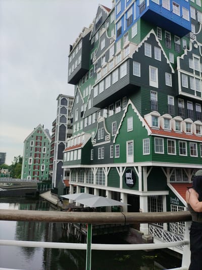 This media file depicts the Inntel Hotels in Zaandam, Netherlands, a uniquely designed hotel with a facade resembling a stack of traditional Dutch houses. The building's exterior is a vibrant mix of green, blue, and gray, with white trim and numerous windows. Several sections of the hotel jut out at different angles, creating a striking and unconventional architectural profile. The hotel is situated along a canal, and the reflection of the building can be seen in the dark water.
In the foreground, a wooden railing stretches across the frame, suggesting the viewer is standing on a bridge or walkway overlooking the canal. A white metal railing is visible beneath the wooden one. To the right of the frame, a person's back is visible, wearing a dark garment and a green cap, and facing away from the camera.
Beneath the hotel's green facade, a sign reads "Inntel hotels." Another sign near the canal level says "Food & Drinks." Umbrellas are set up outside a section of the hotel, hinting at an outdoor dining area. The sky is overcast, suggesting a cloudy or possibly rainy day. The overall scene conveys a sense of urban exploration and appreciation for unique architecture in a waterside setting. The location cues, such as the architectural style and the canal, clearly identify it as Zaandam, Netherlands.