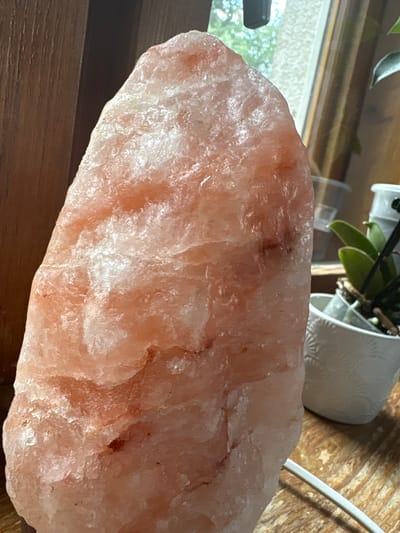 A Himalayan salt lamp, predominantly pink and white, is positioned on a wooden surface. Its rough, crystalline texture is visible. To the right of the lamp, a small potted plant with green leaves in a white, patterned pot is partially visible. Behind the plant, a window with greenery outside can be seen. A white electrical cord extends from the bottom of the lamp towards the right side of the frame. The scene is illuminated by natural light filtering through the window. There are no people or animals present in the image. The specific location in Hradec-Nová Ves, Czechia, cannot be determined from the visual information provided.