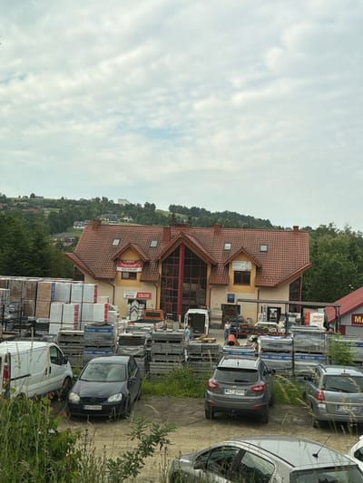 This is an eye-level, outdoor, medium shot captured on a cloudy day. In the background, a two-story building with a red tiled roof, featuring dormer windows and a large multi-paned windowed section on the right, is visible. A red banner reading "LOKALE DO WYNAJECIA" (Premises for Rent) and a phone number is attached to the building. The building is surrounded by a storage area with stacks of construction materials, including pallets of tiles, stone slabs, and white-wrapped items. Several vehicles are parked in the foreground and mid-ground. These include a white van, a dark grey Volkswagen Golf with license plate "KNT 93903", a grey Hyundai SUV with license plate "KL 47693", a silver Opel Astra with license plate "KL 4428", and a partial view of a silver car in the lower foreground. A small orange truck is also visible among the stored materials. The ground is unpaved, with dirt and sparse vegetation. The scene appears to be a commercial property, possibly a building supply store or a business complex, located in Limanowa, Poland. The environment is hilly with trees and houses visible on the distant slopes.