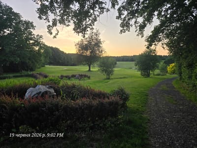 The image depicts a serene golf course at dusk, bathed in the warm glow of the setting sun. The foreground is dominated by lush, dark green bushes and a large, grey boulder. A gravel path curves its way through the scene, leading the eye towards the gently rolling green hills of the golf course in the background. Scattered trees and manicured fairways are visible, punctuated by a sand trap. The sky is a pale gradient of orange and yellow, transitioning to a soft grey, with silhouetted trees forming a natural frame around the upper portion of the image. A timestamp in the lower left corner reads "19 червня 2026 р. 9:02 PM". While the exact location is not explicitly stated in the image, the timestamp, which appears to be in Ukrainian, suggests a potential connection to Ukraine or a Ukrainian speaker. However, the prompt specifies the location as Bannewitz, Germany, which is not visually corroborated by the image content. There are no people or animals visible in the scene. The overall atmosphere is peaceful and tranquil, characteristic of a late evening on a golf course.
